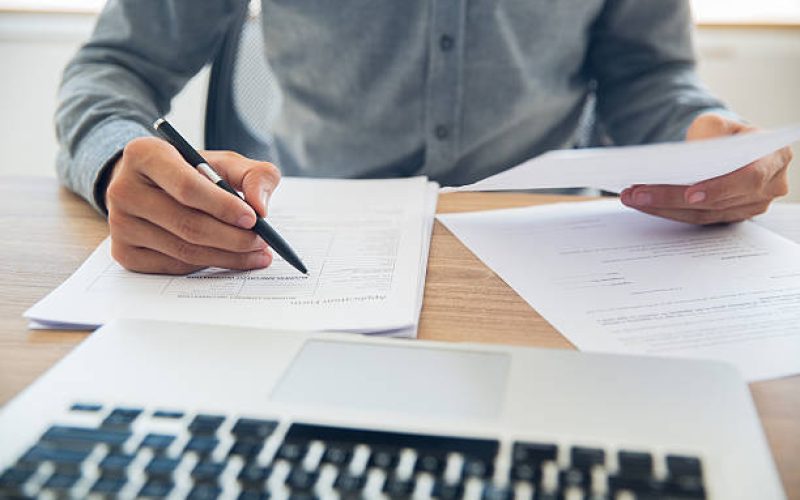 Unrecognizable businessman sitting at table holding ballpoint pen and papers and checking documents in office
